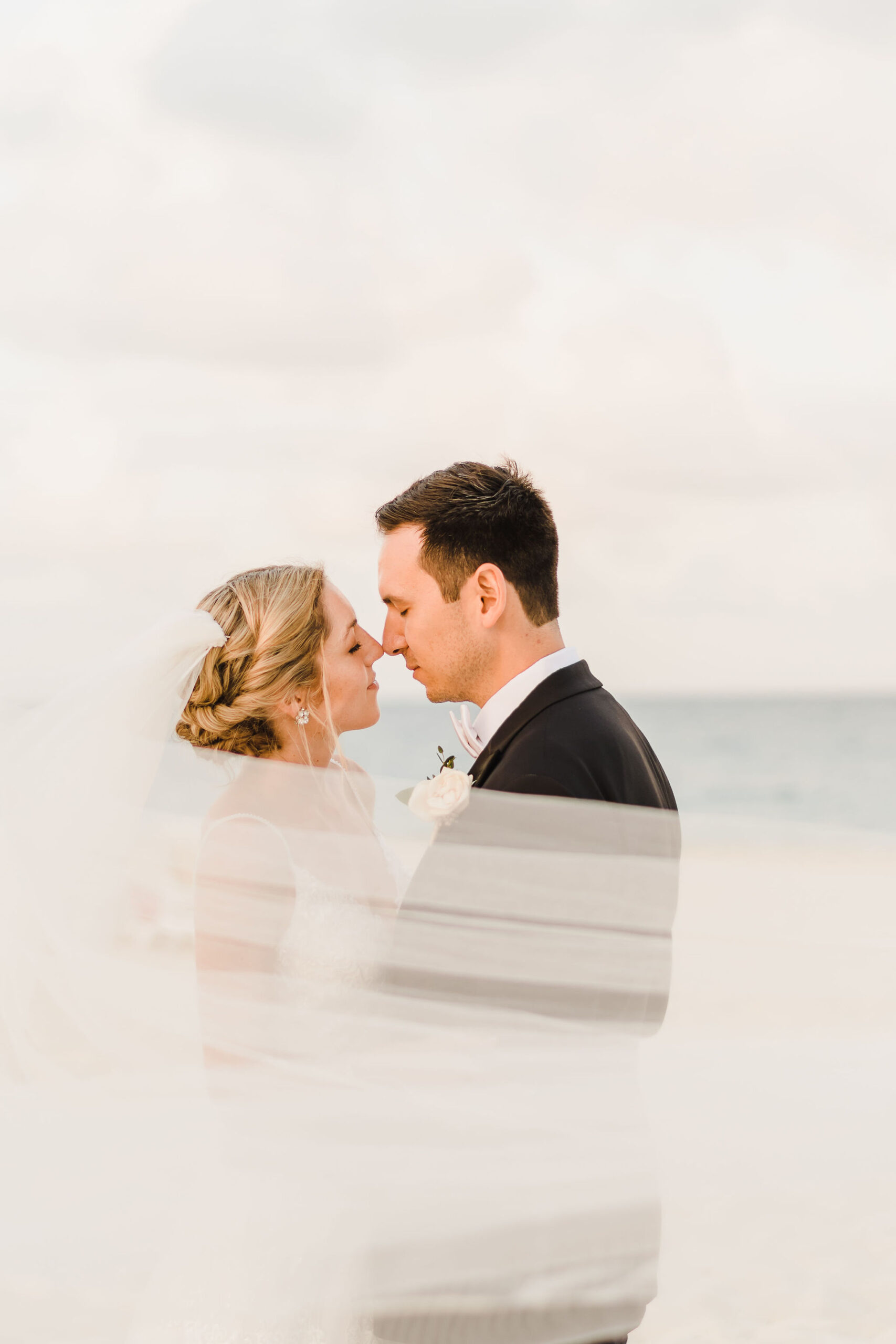bride and groom embrace on a beach while veil swoops across them.