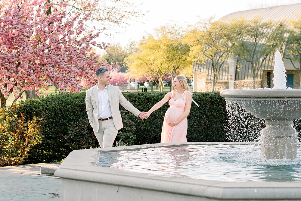 A pregnant woman wearing a pink dress holds her husband's hand during their Baker Park maternity photos in Frederick, Maryland.