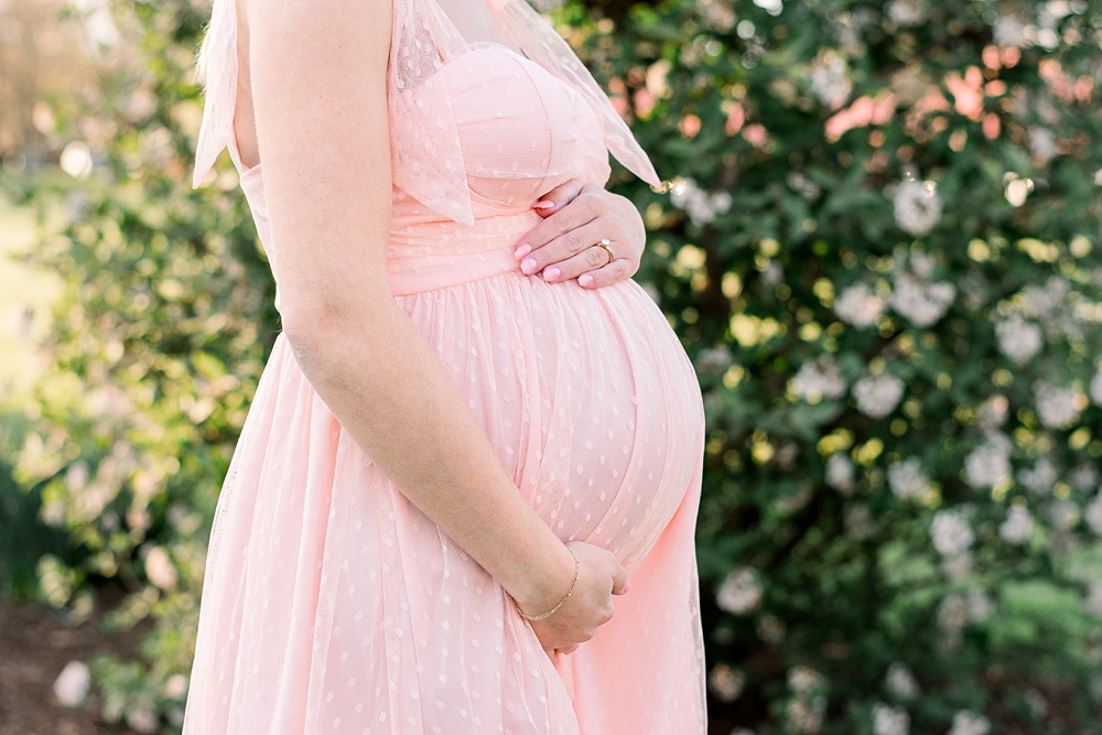 A pregnant woman wearing a pink dress holds her belly during her Baker Park maternity photos in Frederick, Maryland.