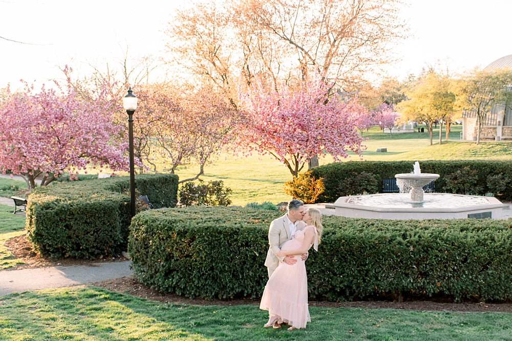 A pregnant woman wearing a pink dress embraces her husband during their Baker Park maternity photos in Frederick, Maryland.