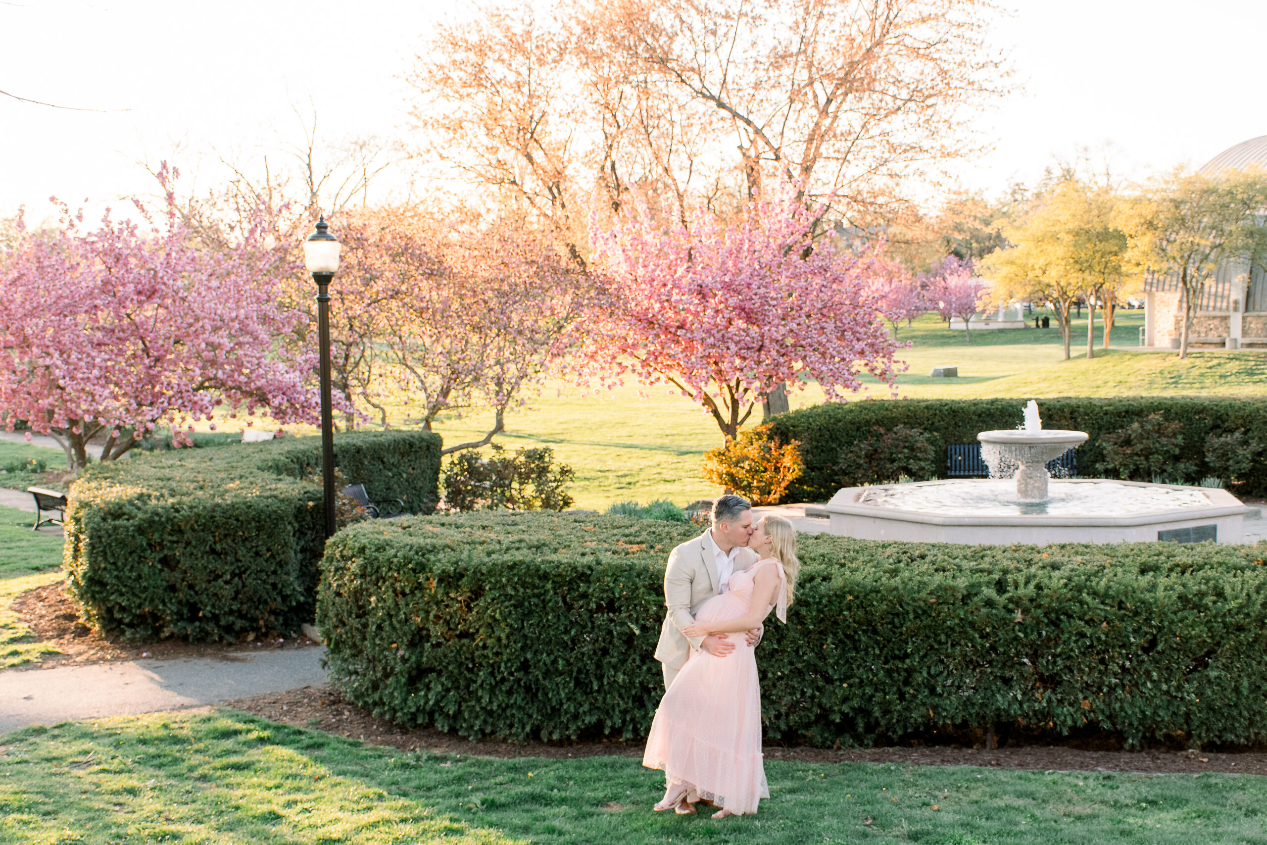 A pregnant woman wearing a pink dress embraces her husband during their Baker Park maternity photos in Frederick, Maryland.