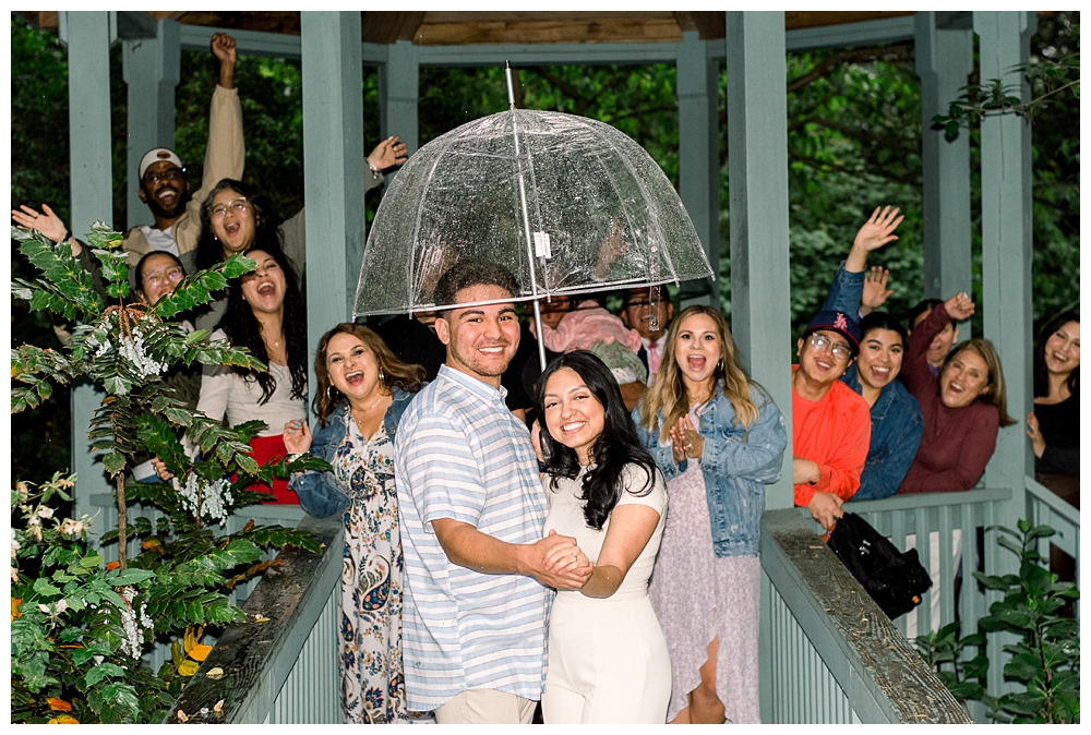 A group of friends and family stand with a recently engaged couple after a surprise proposal at McCrillis Gardens in Bethesda, Maryland.