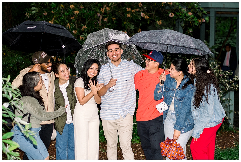 A group of friends and family stand with a recently engaged couple after a surprise proposal at McCrillis Gardens in Bethesda, Maryland.