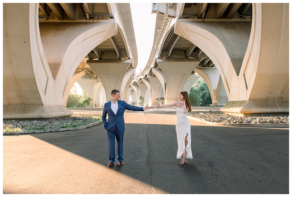 A couple stands together underneath the Woodrow Wilson Bridge at Jones Point Park in Old Town Alexandria, Virginia, during their engagement session.